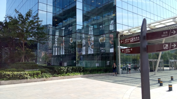 A modern glass building with reflective windows stands prominent in an urban area, surrounded by lush greenery. There are signs in the foreground providing directions for a subway, dining area, and bicycle parking. People are visible walking near the entrance, contributing to the active atmosphere.