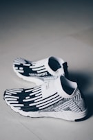 A pair of black and white slip-on sneakers with a modern, knitted design featuring horizontal and vertical patterns. The sole of the shoes is white with black accents. The background is a neutral, light-colored floor, creating a minimalist aesthetic.