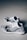 A pair of black and white slip-on sneakers with a modern, knitted design featuring horizontal and vertical patterns. The sole of the shoes is white with black accents. The background is a neutral, light-colored floor, creating a minimalist aesthetic.