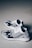 A pair of black and white slip-on sneakers with a modern, knitted design featuring horizontal and vertical patterns. The sole of the shoes is white with black accents. The background is a neutral, light-colored floor, creating a minimalist aesthetic.
