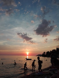 Children are playing in the shallow water of a beach during sunset. The sky is filled with scattered clouds, and the sun is setting on the horizon, casting a warm glow over the sea. Silhouettes of trees and people add to the serene and playful atmosphere.