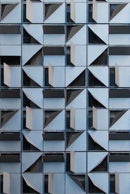 A grid of architectural panels arranged in a repetitive geometric pattern featuring angular, folded elements. Each panel appears to be a section of a building façade with openings and overlapping metal sheets, creating an abstract, three-dimensional effect.