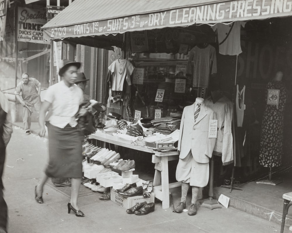 grayscale photo of woman walking in front of shoe store