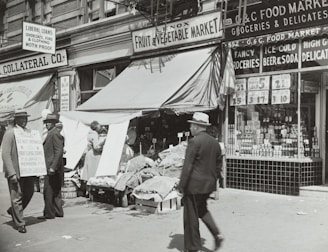 A black-and-white street scene featuring a fruit and vegetable market with an awning, surrounded by various storefronts. Two men in suits and hats stand to the left, one holding a sign with text. Another man walks by in the foreground. Various goods and advertisements are visible in the shop windows, with prices prominently displayed.