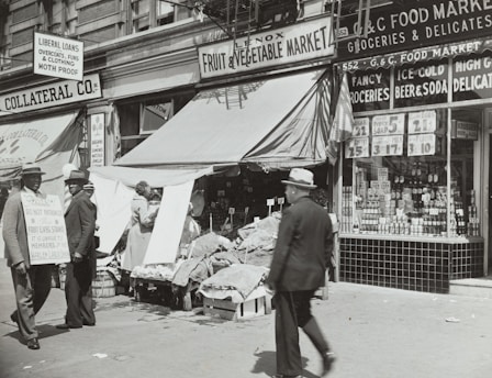 A black-and-white street scene featuring a fruit and vegetable market with an awning, surrounded by various storefronts. Two men in suits and hats stand to the left, one holding a sign with text. Another man walks by in the foreground. Various goods and advertisements are visible in the shop windows, with prices prominently displayed.