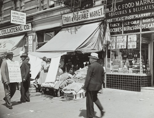 A black-and-white street scene featuring a fruit and vegetable market with an awning, surrounded by various storefronts. Two men in suits and hats stand to the left, one holding a sign with text. Another man walks by in the foreground. Various goods and advertisements are visible in the shop windows, with prices prominently displayed.