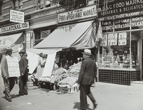 A black-and-white street scene featuring a fruit and vegetable market with an awning, surrounded by various storefronts. Two men in suits and hats stand to the left, one holding a sign with text. Another man walks by in the foreground. Various goods and advertisements are visible in the shop windows, with prices prominently displayed.