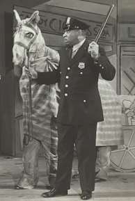 An actor in police uniform standing beside a vintage police cruiser on location.