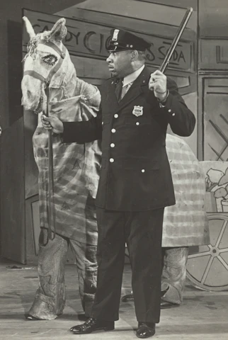 An actor in police uniform standing beside a vintage police cruiser on location.