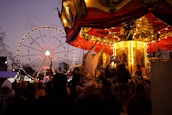 A lively carnival scene featuring a carousel with bright, warm lights and intricate details. In the background, a large Ferris wheel is illuminated against the evening sky. Several people are gathered, enjoying the festive atmosphere.