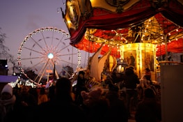 A lively carnival scene featuring a carousel with bright, warm lights and intricate details. In the background, a large Ferris wheel is illuminated against the evening sky. Several people are gathered, enjoying the festive atmosphere.