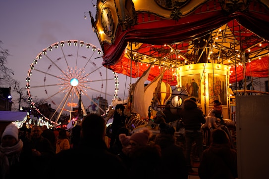 A lively carnival scene featuring a carousel with bright, warm lights and intricate details. In the background, a large Ferris wheel is illuminated against the evening sky. Several people are gathered, enjoying the festive atmosphere.