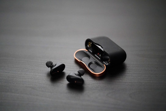 A pair of wireless earbuds with a sleek design are placed on a dark surface. The earbuds are next to an open charging case, which has a metallic accent around the edges. The surface has a textured appearance, adding a minimalist and modern aesthetic.