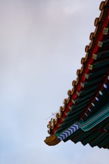A traditional architectural roof corner with intricate details, featuring red, green, and gold elements against a cloudy sky.