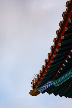 A traditional architectural roof corner with intricate details, featuring red, green, and gold elements against a cloudy sky.