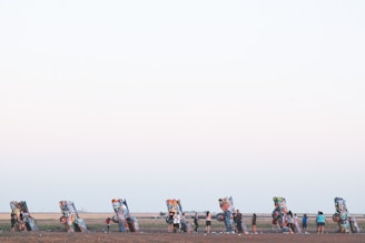 A row of brightly painted, partially buried cars stand out against a flat, expansive landscape. Several people are interacting with these car sculptures, adding their own graffiti or examining the artwork. The open field has a vast sky above, hinting at a serene and open environment.