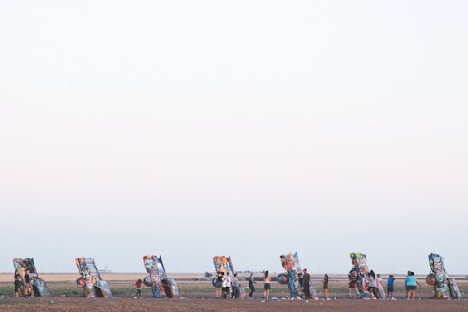 A row of brightly painted, partially buried cars stand out against a flat, expansive landscape. Several people are interacting with these car sculptures, adding their own graffiti or examining the artwork. The open field has a vast sky above, hinting at a serene and open environment.