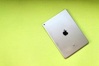 A silver iPad with the Apple logo on the back rests on a bright lime green background. The iPad is positioned diagonally, showcasing its sleek metallic design and minimalistic aesthetic.