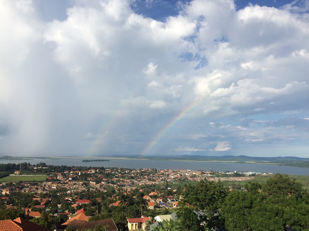 aerial photography of houses on green field viewing body of water and mountain with rainbow under white and blue sky