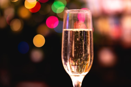 A tall, elegant glass filled with sparkling champagne set against a backdrop of colorful bokeh lights. The soft focus of the background emphasizes the clarity and effervescence of the drink, creating a festive and lively atmosphere.