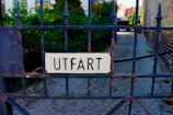 A wrought iron gate with a sign that reads 'UTFART', which is attached in the center. The background includes some greenery and an alleyway with gravel on the ground.