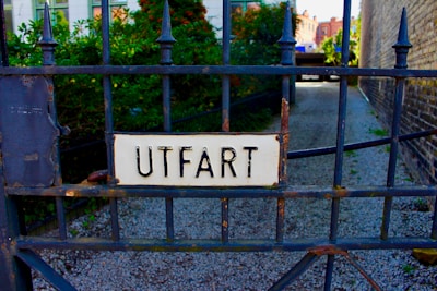 A wrought iron gate with a sign that reads 'UTFART', which is attached in the center. The background includes some greenery and an alleyway with gravel on the ground.