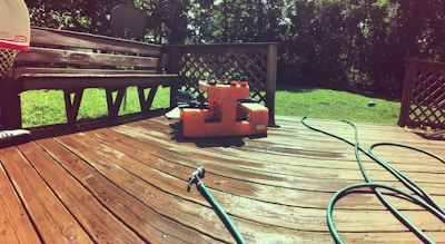 A wooden deck is illuminated by sunlight, showing a few garden hoses laying across it. An orange water pump or utility box is placed centrally on the deck near a bench. The background features green grass and trees, with a satellite dish visible.