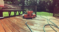 A wooden deck is illuminated by sunlight, showing a few garden hoses laying across it. An orange water pump or utility box is placed centrally on the deck near a bench. The background features green grass and trees, with a satellite dish visible.