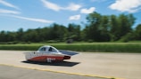 A streamlined solar-powered car is driving on a road with a lush green forest backdrop. The car features a sleek aerodynamic design with solar panels on its surface and carries the number 92 along with various logos. The background is slightly blurred, suggesting high speed.