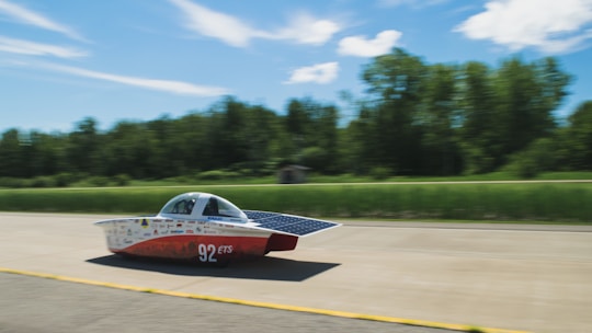 A streamlined solar-powered car is driving on a road with a lush green forest backdrop. The car features a sleek aerodynamic design with solar panels on its surface and carries the number 92 along with various logos. The background is slightly blurred, suggesting high speed.