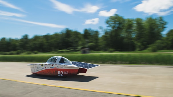 A streamlined solar-powered car is driving on a road with a lush green forest backdrop. The car features a sleek aerodynamic design with solar panels on its surface and carries the number 92 along with various logos. The background is slightly blurred, suggesting high speed.