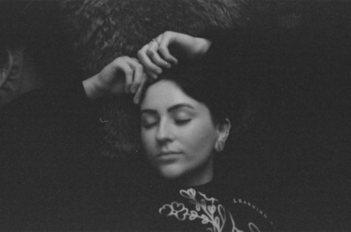 A black and white photograph features a person lying down with eyes closed. The gentle expression suggests a state of relaxation. Hands are positioned near the head, and there's a decorative pattern visible on the clothing.