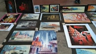 Several paintings are laid out on the ground, displaying various scenes with vibrant colors. These include depictions of buildings, landscapes, a boat on water, and a person in traditional attire. The artworks are arranged on a pavement, held down by stones at the corners.