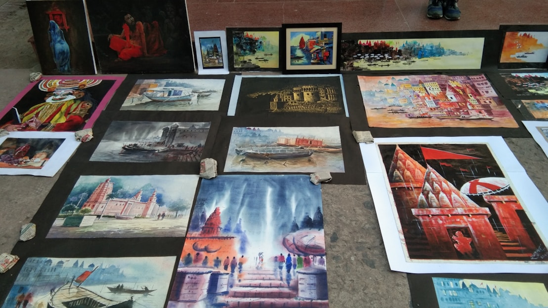 Several paintings are laid out on the ground, displaying various scenes with vibrant colors. These include depictions of buildings, landscapes, a boat on water, and a person in traditional attire. The artworks are arranged on a pavement, held down by stones at the corners.