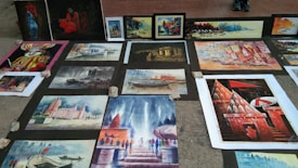 Several paintings are laid out on the ground, displaying various scenes with vibrant colors. These include depictions of buildings, landscapes, a boat on water, and a person in traditional attire. The artworks are arranged on a pavement, held down by stones at the corners.