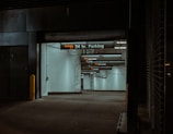 A dimly lit parking garage entrance with a sign indicating '24 hr. Parking'. The walls are tiled in white, and several overhead signs provide instructions like 'pull ahead' and 'drive slowly'. The atmosphere is somewhat shadowy, with industrial elements such as pipes visible.
