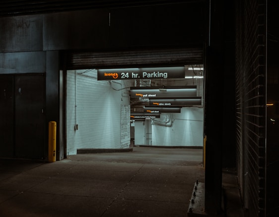 A dimly lit parking garage entrance with a sign indicating '24 hr. Parking'. The walls are tiled in white, and several overhead signs provide instructions like 'pull ahead' and 'drive slowly'. The atmosphere is somewhat shadowy, with industrial elements such as pipes visible.