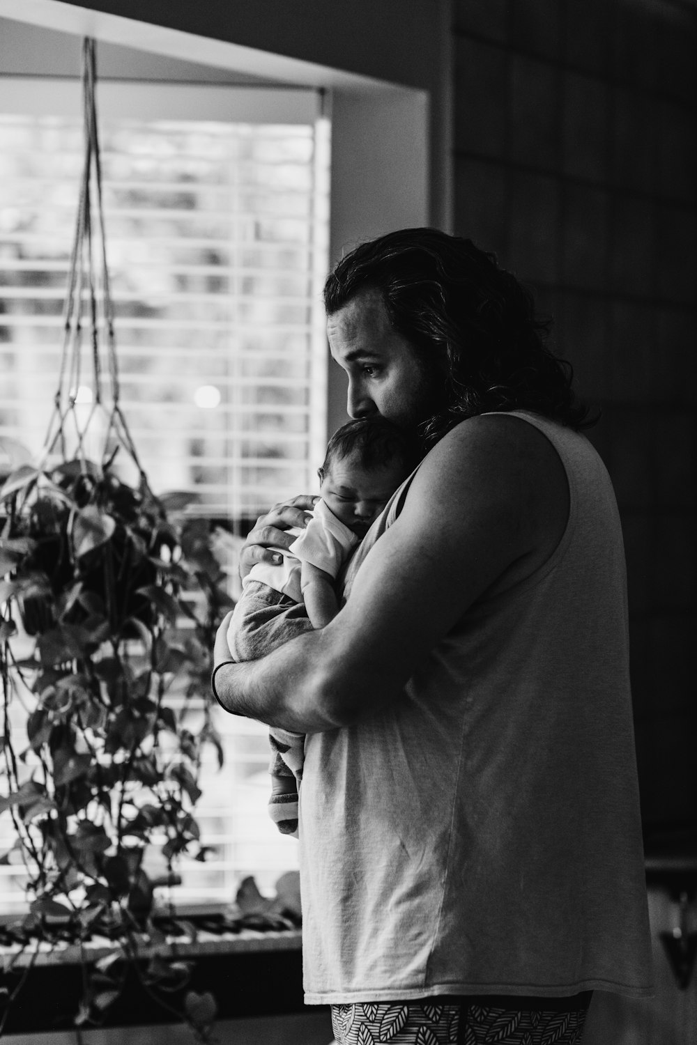 grayscale photography of man wearing tank top carrying newborn baby while standing near window