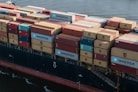 A large cargo ship loaded with numerous shipping containers stacked in various colors and brands, floating on a body of water. The containers display logos like MAERSK and MSC. The vessel's hull is visible, painted in a dark color with a visible watermark on its side.