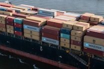 A large cargo ship loaded with numerous shipping containers stacked in various colors and brands, floating on a body of water. The containers display logos like MAERSK and MSC. The vessel's hull is visible, painted in a dark color with a visible watermark on its side.