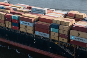 A large cargo ship loaded with numerous shipping containers stacked in various colors and brands, floating on a body of water. The containers display logos like MAERSK and MSC. The vessel's hull is visible, painted in a dark color with a visible watermark on its side.