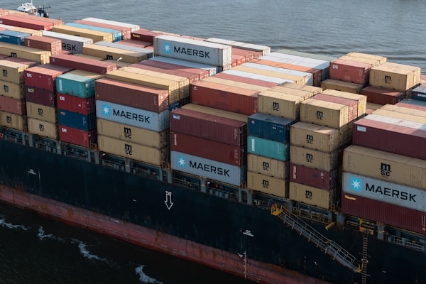 A large cargo ship loaded with numerous shipping containers stacked in various colors and brands, floating on a body of water. The containers display logos like MAERSK and MSC. The vessel's hull is visible, painted in a dark color with a visible watermark on its side.