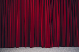 Richly textured red velvet theatre curtains hanging in vertical folds, hinting at a stage beyond. The fabric appears heavy and luxurious, with a subtle sheen.