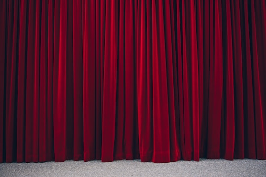 Richly textured red velvet theatre curtains hanging in vertical folds, hinting at a stage beyond. The fabric appears heavy and luxurious, with a subtle sheen.