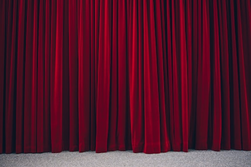Richly textured red velvet theatre curtains hanging in vertical folds, hinting at a stage beyond. The fabric appears heavy and luxurious, with a subtle sheen.