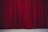Richly textured red velvet theatre curtains hanging in vertical folds, hinting at a stage beyond. The fabric appears heavy and luxurious, with a subtle sheen.