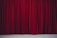 Richly textured red velvet theatre curtains hanging in vertical folds, hinting at a stage beyond. The fabric appears heavy and luxurious, with a subtle sheen.