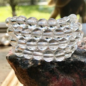 A stack of clear glass beads is artistically arranged on a textured stone surface. The sunlight reflects off the beads, highlighting their shiny and smooth surfaces. The background is softly blurred with a suggestion of green foliage, adding a natural element to the composition.