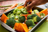 A hand is placing broccoli florets into a metal baking tray filled with vibrant, fresh vegetables including chunks of orange pumpkin and slices of carrot. More carrots and asparagus can be seen in the background on a speckled countertop.