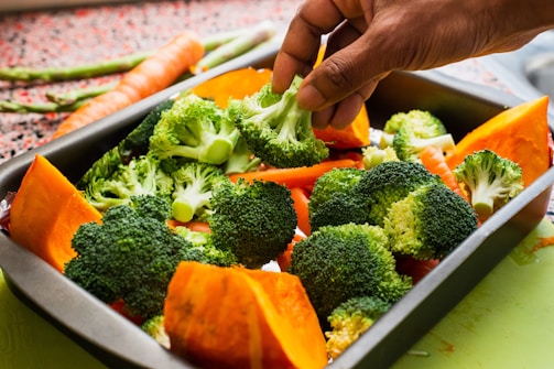 A hand is placing broccoli florets into a metal baking tray filled with vibrant, fresh vegetables including chunks of orange pumpkin and slices of carrot. More carrots and asparagus can be seen in the background on a speckled countertop.